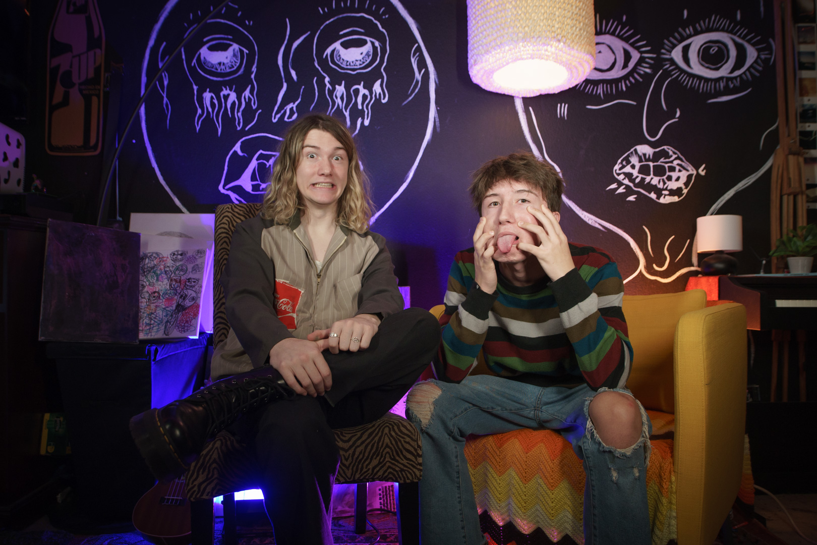 A man with long blond hair named Parker James sits next to his brother, Caden Shea in front of a mural of abstract faces Musicians and Parker James sit in front of a mural of abstract faces .