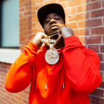 Rapper Benny the Butcher, wearing a red shirt, faces the camera and is holding up his gold chains
