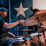 A drummer plays drums on stage in front of a big Texas flag.