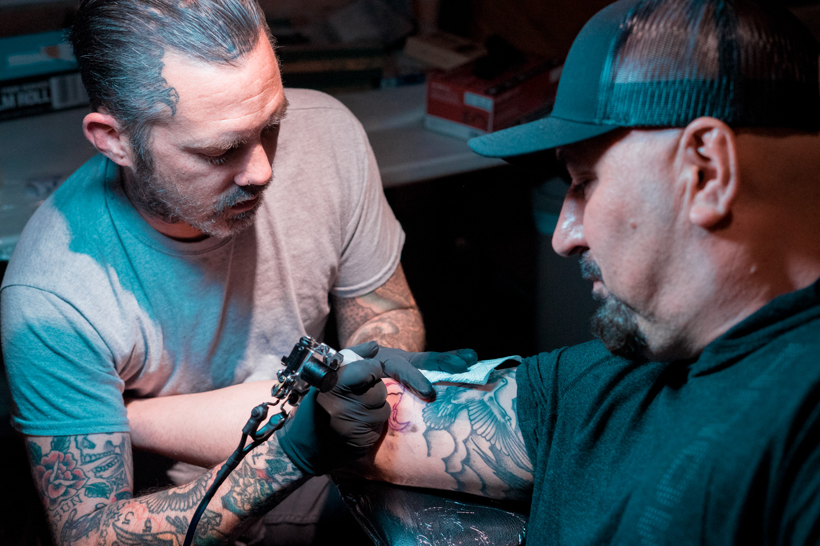 A man applies a bull skull tattoo to another man's inner right bicep.