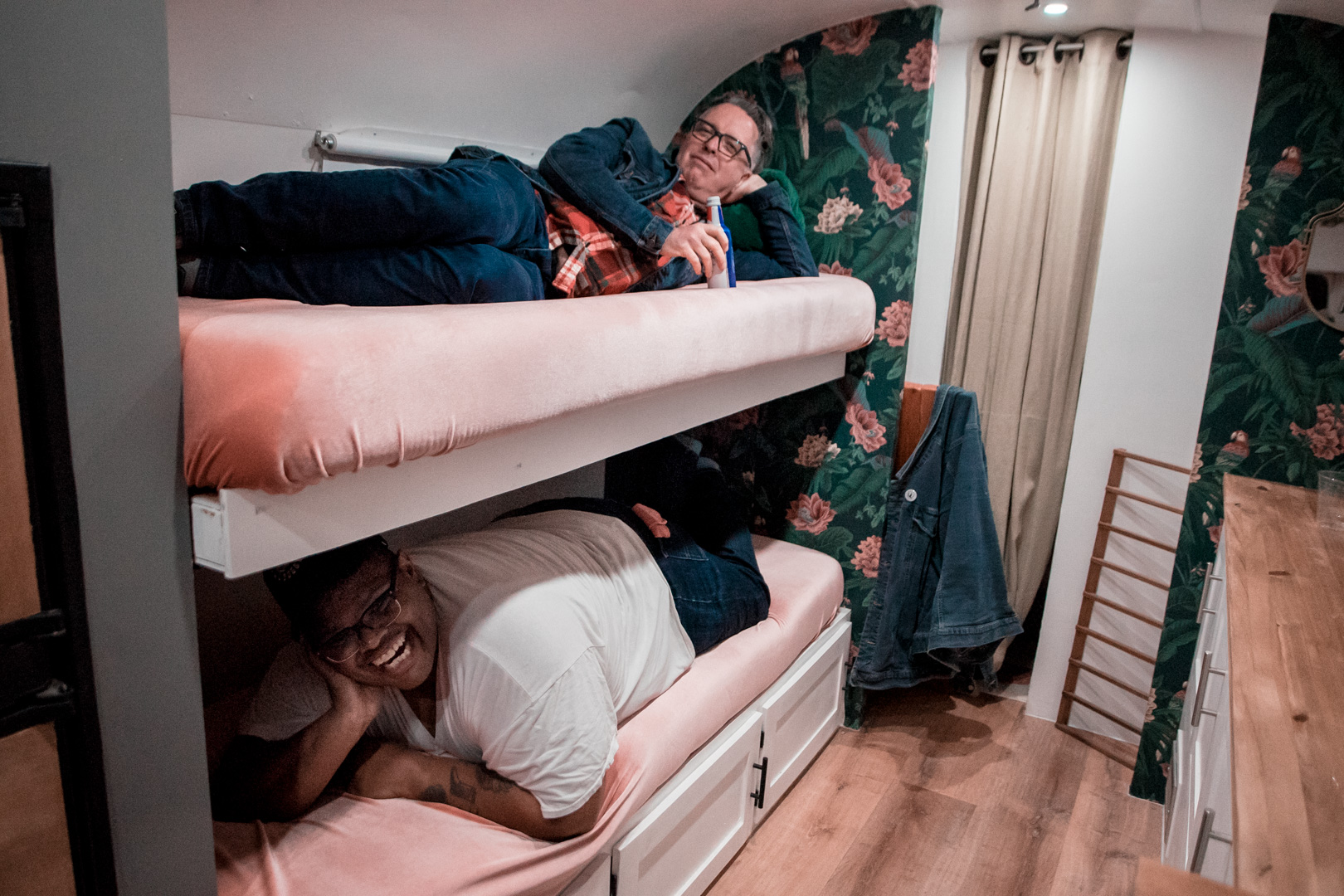 Two men lay on plush pink bunk beds inside of an airstream, smiling at the camera.