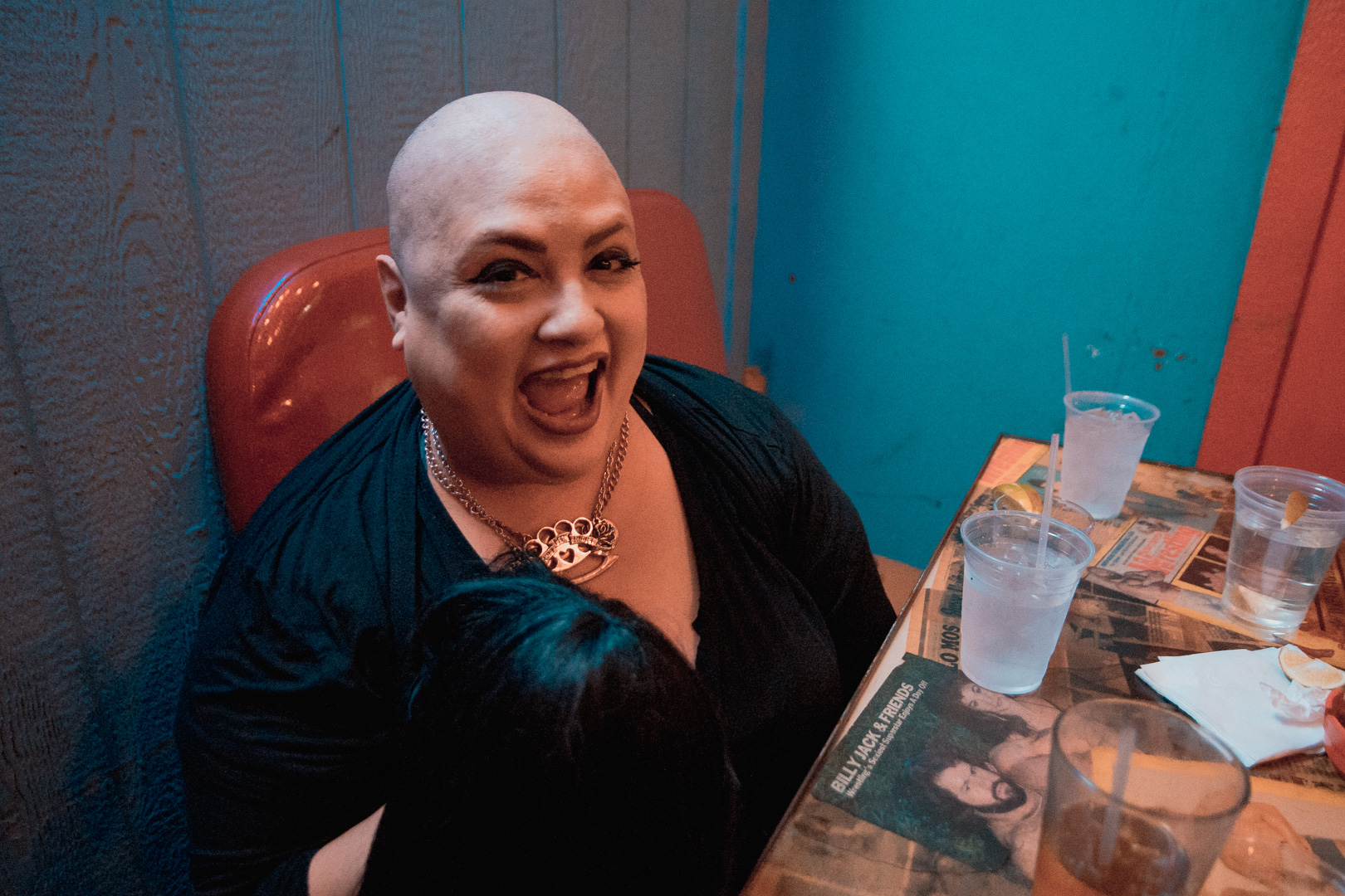 A smiling bald woman sitting at a table holding a black wig. 