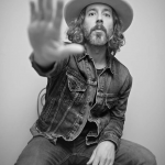 Singer-songwriter Ryan Hamilton is holding his hand out to the camera, palm facing the viewer. He's wearing a jean jacket, jeans and a cowboy hat.