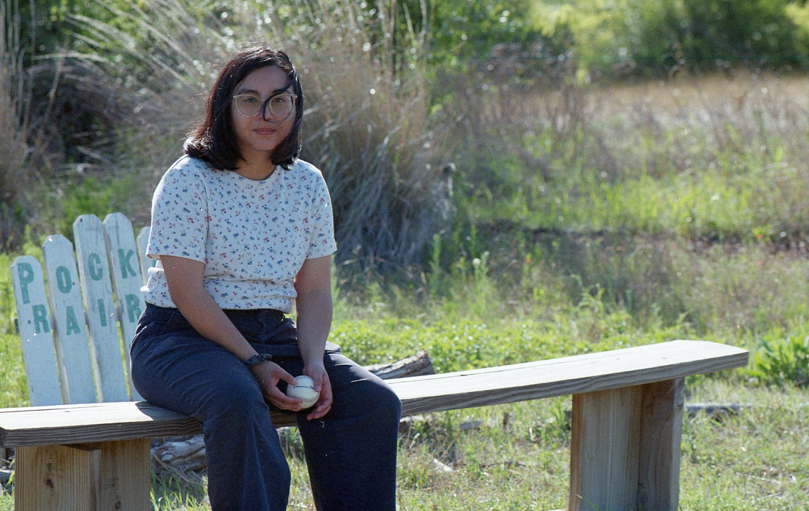 Singer-songwriter Alex Montenegro, who performs as Skirts, sits on a bench at one end on a sunny day, wearing a white shirt and jeans