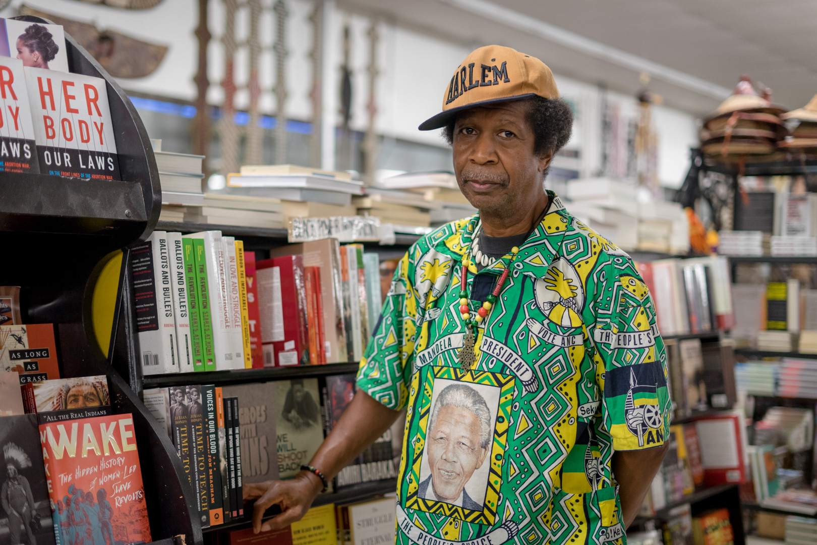 A middle aged man stands next to a bookshelf wearing a Nelson Mandala shirt and Harlem letter hat. 