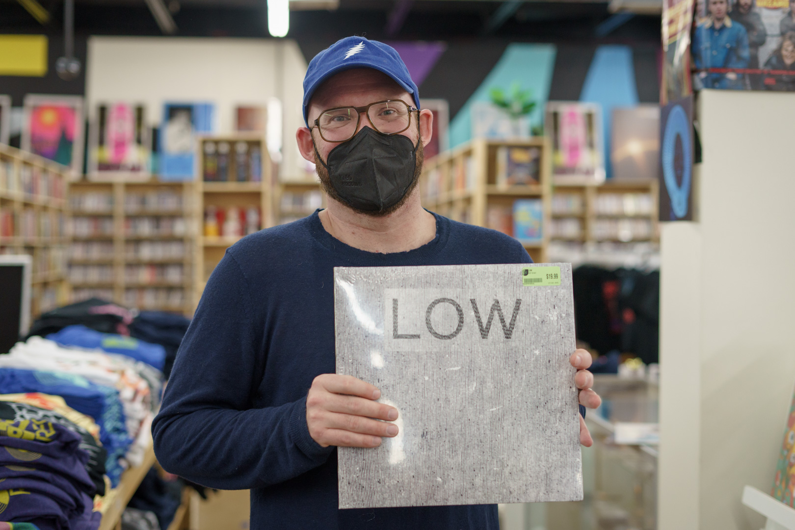 A man in a blue baseball cap and black face mask holds a white vinyl record album with minimalist line artwork.