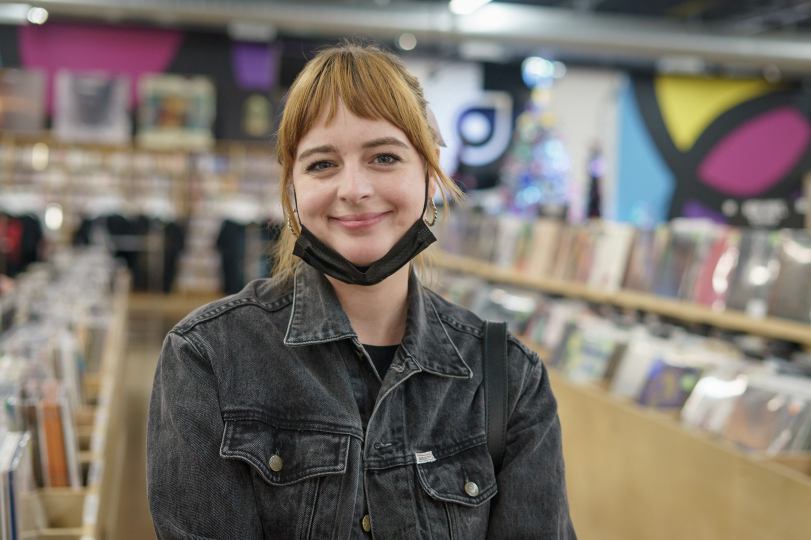A red haired woman in a black denim jacket smiles in a record store.