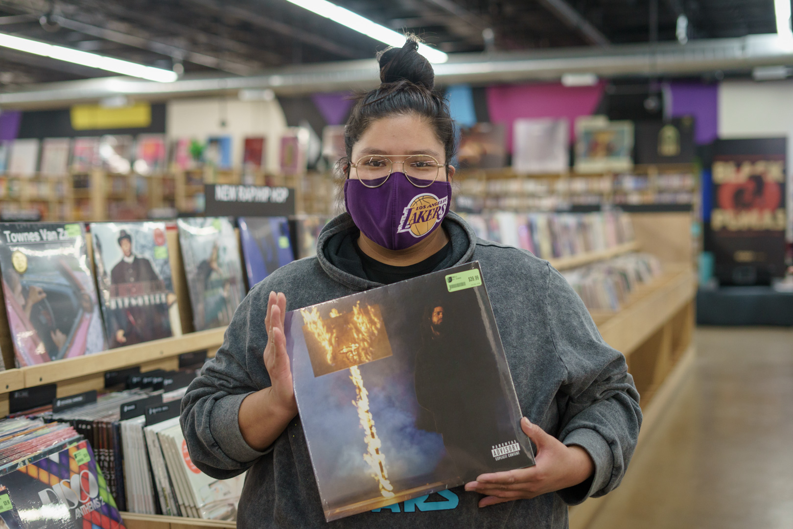 A woman with her hair up in a top bun and wearing purple Lakers face mask holds a vinyl record with a portrait of J. Cole and a street sign on fire. 