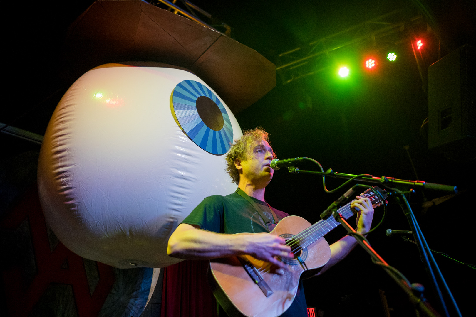 A man plays guitar on stage with a giant eyeball wearing a cowboy hat hangs behind him. 