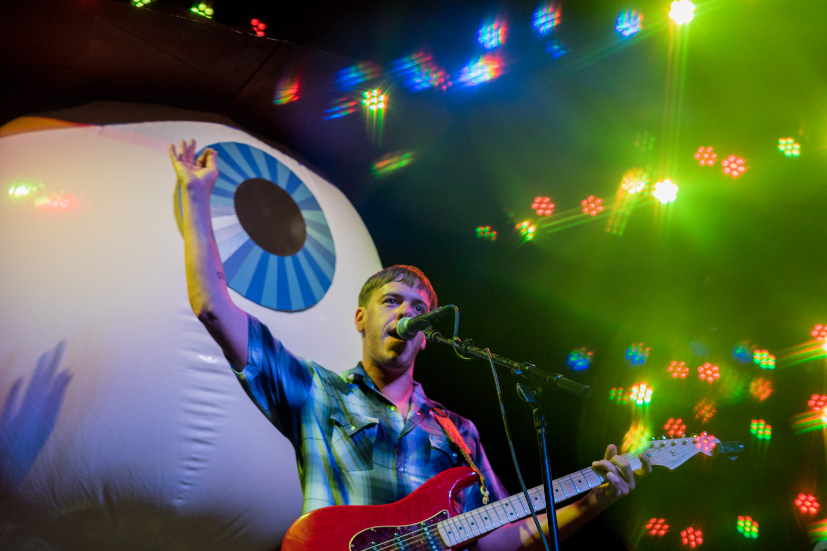 A man on stage playing guitar holds up his hand while a giant eyeball wearing a cowboy hat hangs from the ceiling behind him.
