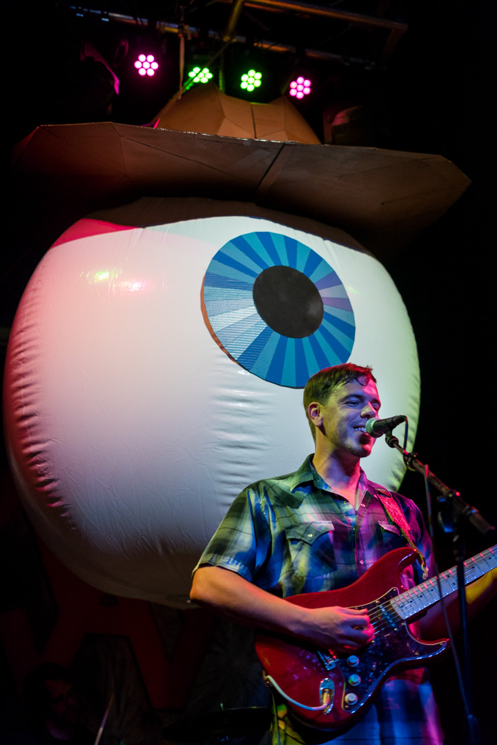 A man smiles on stage in front of giant eyeball with a cowboy hat.