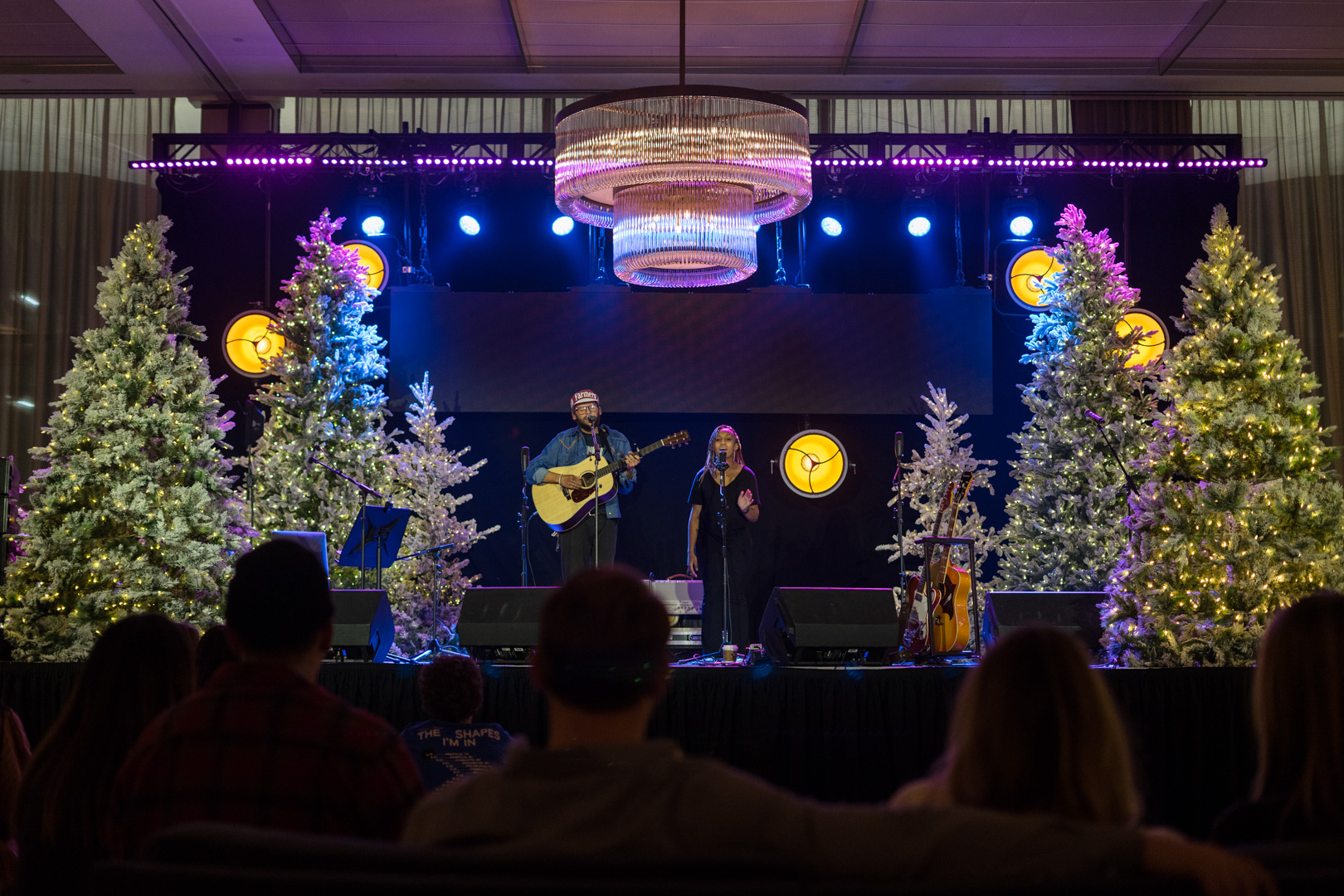 A man plays guitar while singing, a young woman duets with him standing on a stage with large Christmas trees on both sides. 