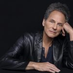 Lindsey Buckingham, wearing a black leather jacket, is staring at the camera and resting his head on one hand