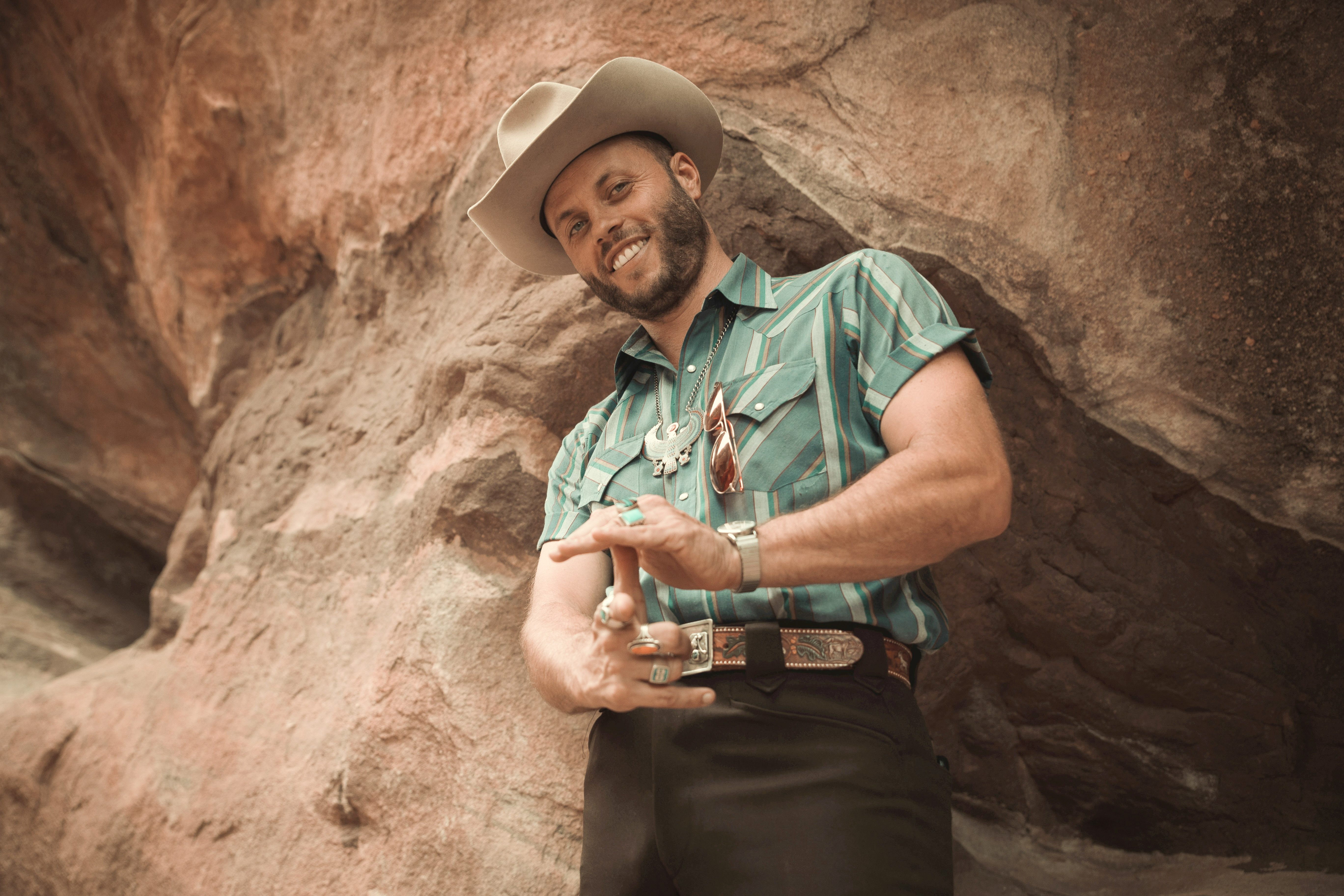A man in a teal and white shirt and a cowboy hat poses for the camera in front of natural rock formation