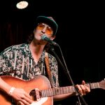 A man in a baseball hat and orange glasses plays guitar on stage and sings.