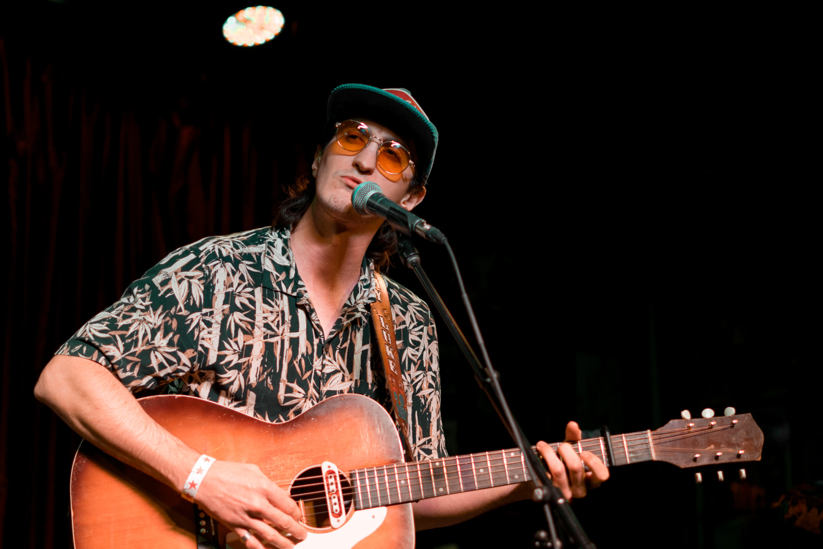 A man in a baseball hat and orange glasses plays guitar on stage and sings. 