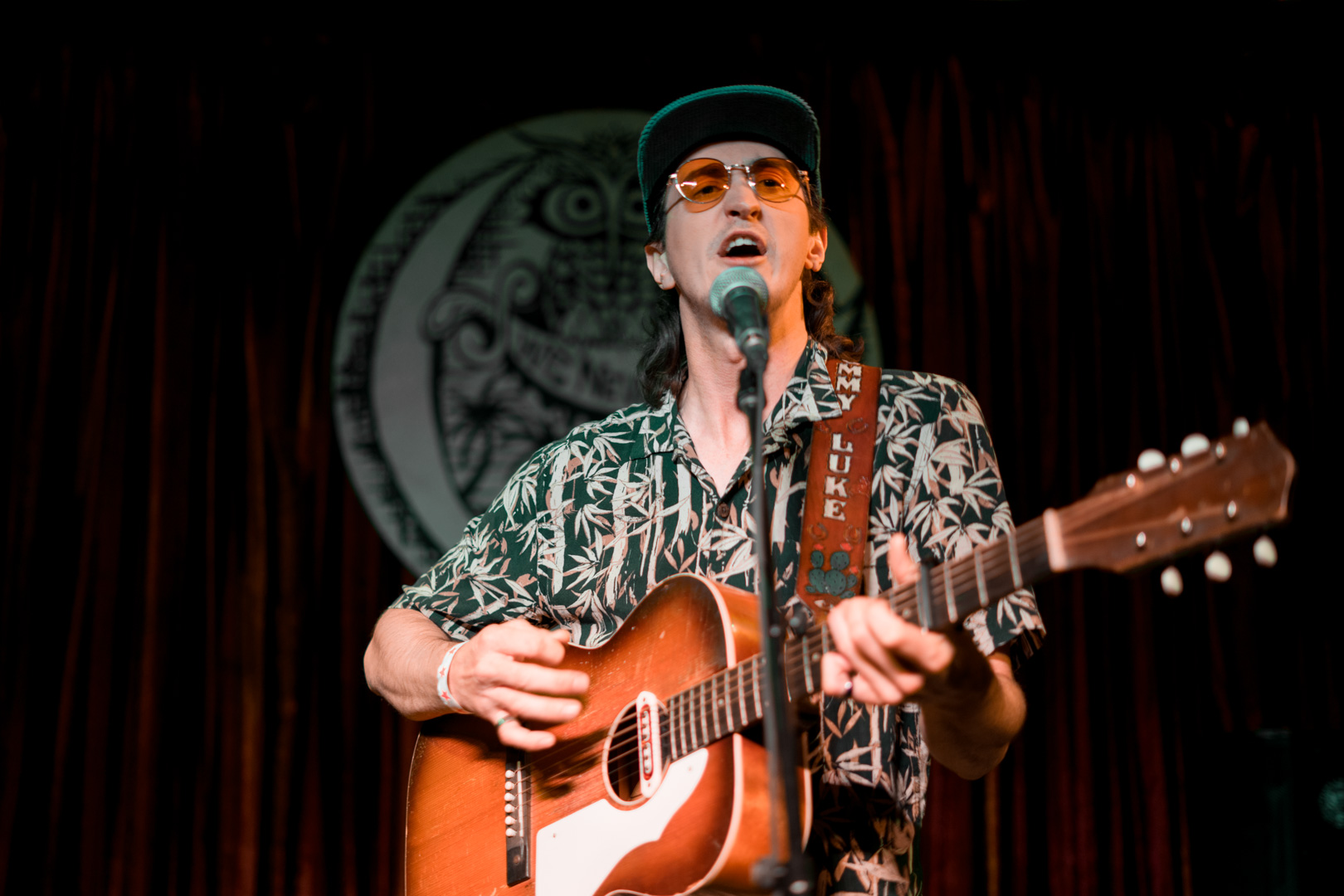 A man in a baseball hat and orange sunglasses plays guitar and sings on stage.