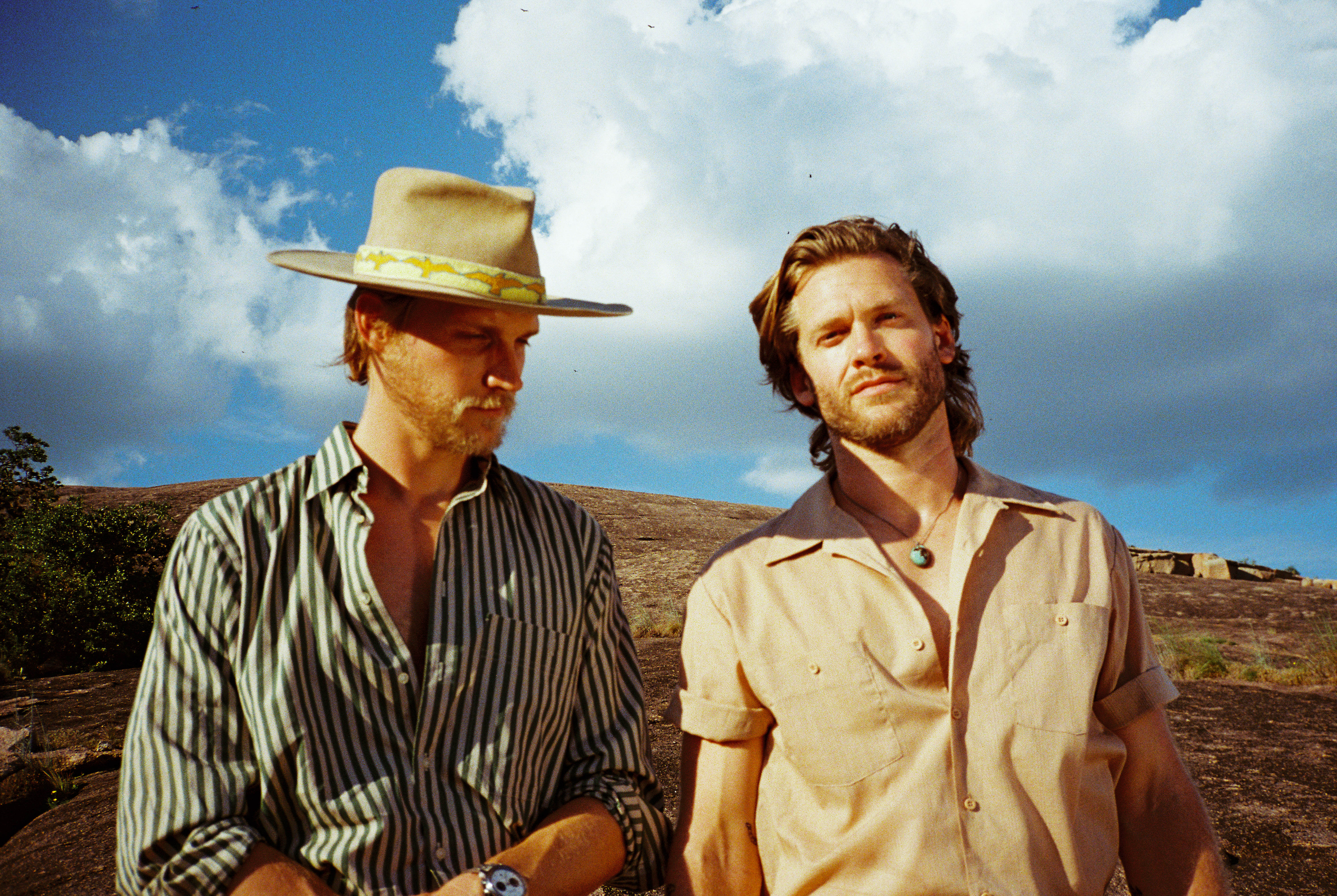 Zach Chance, wearing a hat and a shirt with green stripes, stands next to Jonathan Clay, wearing a tan shirt, outside under a cloudy sky. 