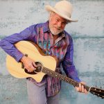 Robert Earl Keen, wearing a cowboy hat and holding an acoustic guitar, faces the camera