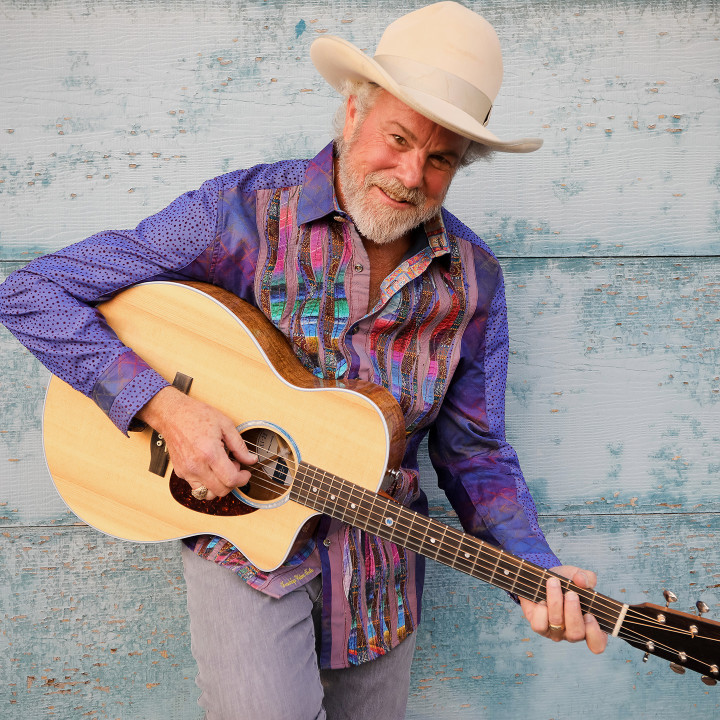 Robert Earl Keen, wearing a cowboy hat and holding an acoustic guitar, faces the camera