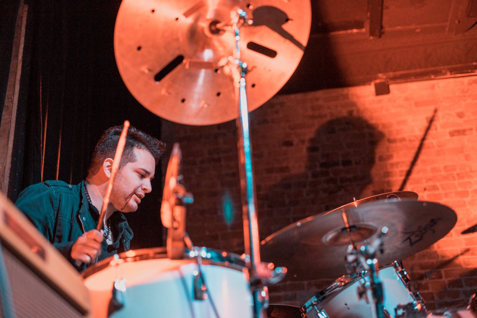 A drummer plays with stage lighting creating a shadow behind him.