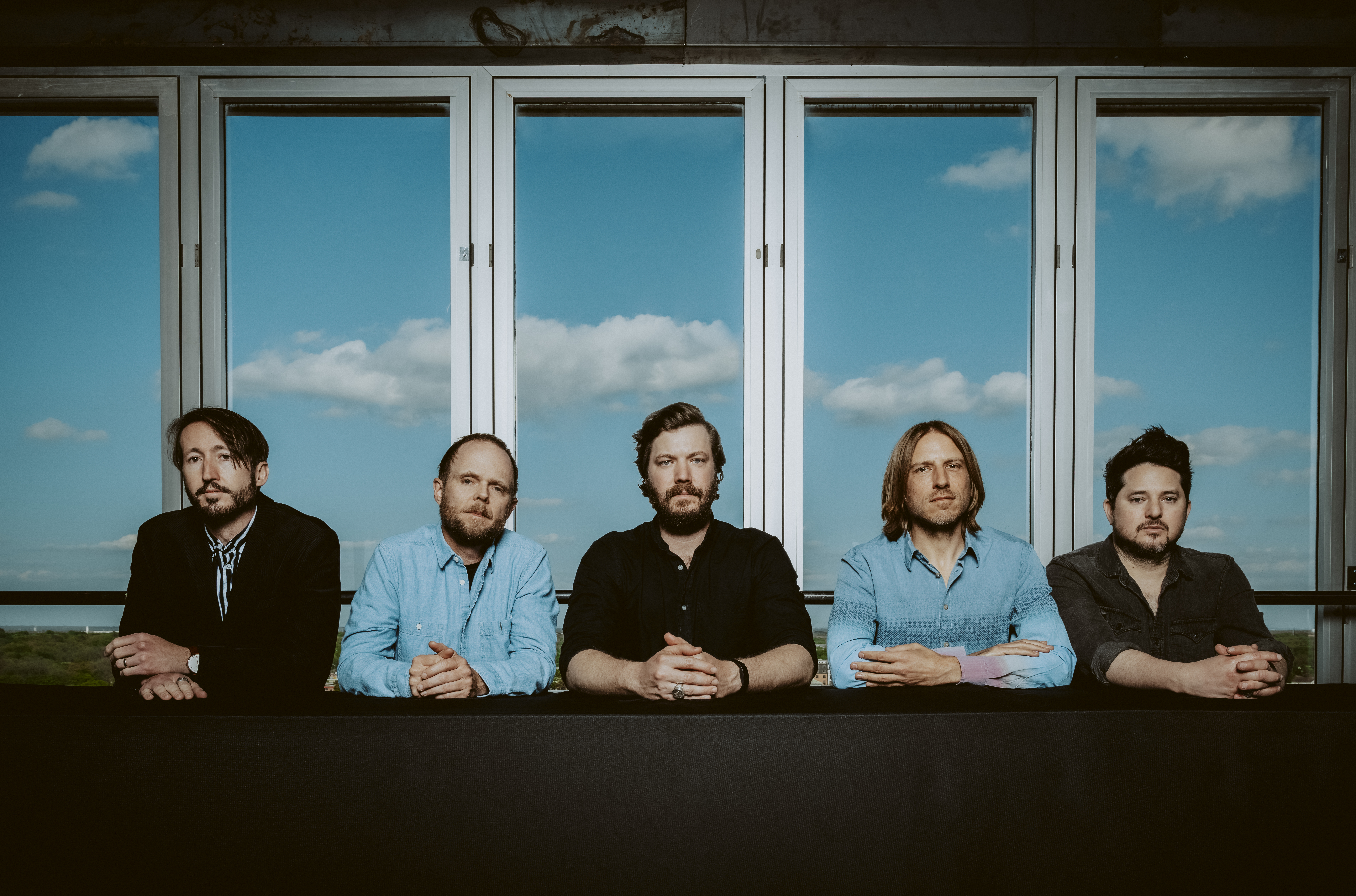 The five members of Midlake are in front of large windows, with blue sky visible