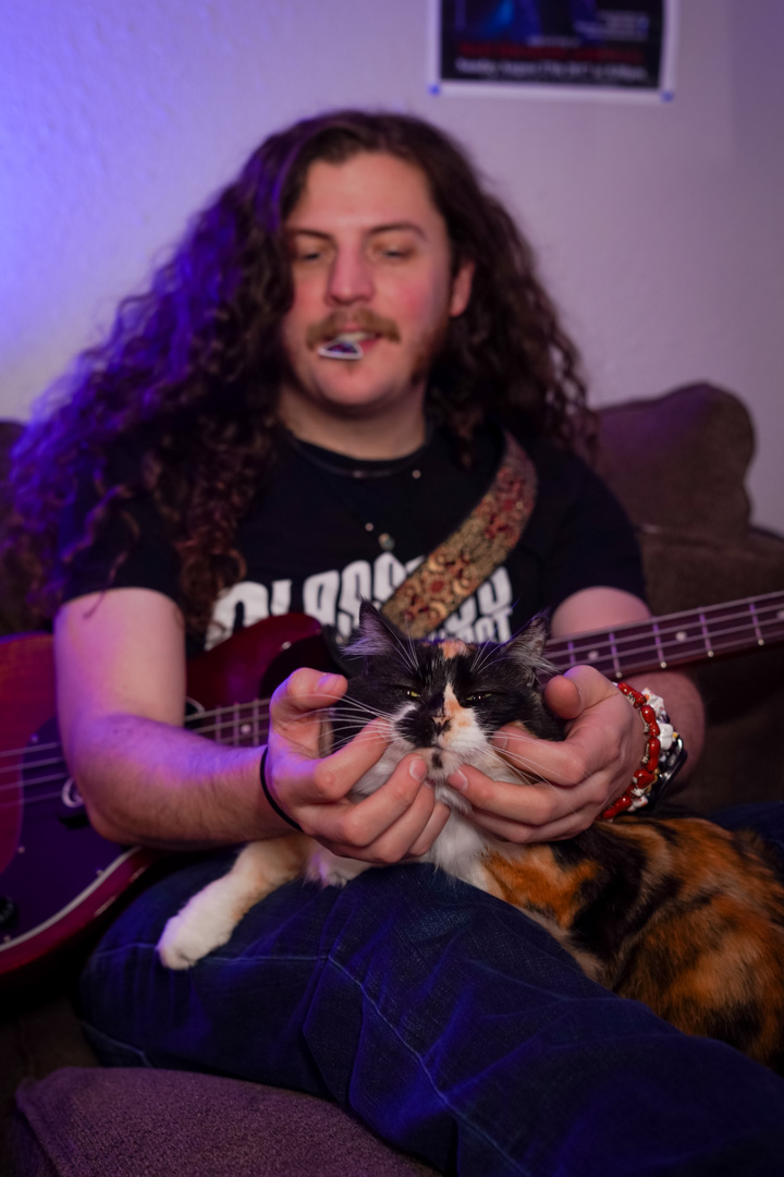 A young man with long hair pets a cat on his lap while a guitar is strapped to him
