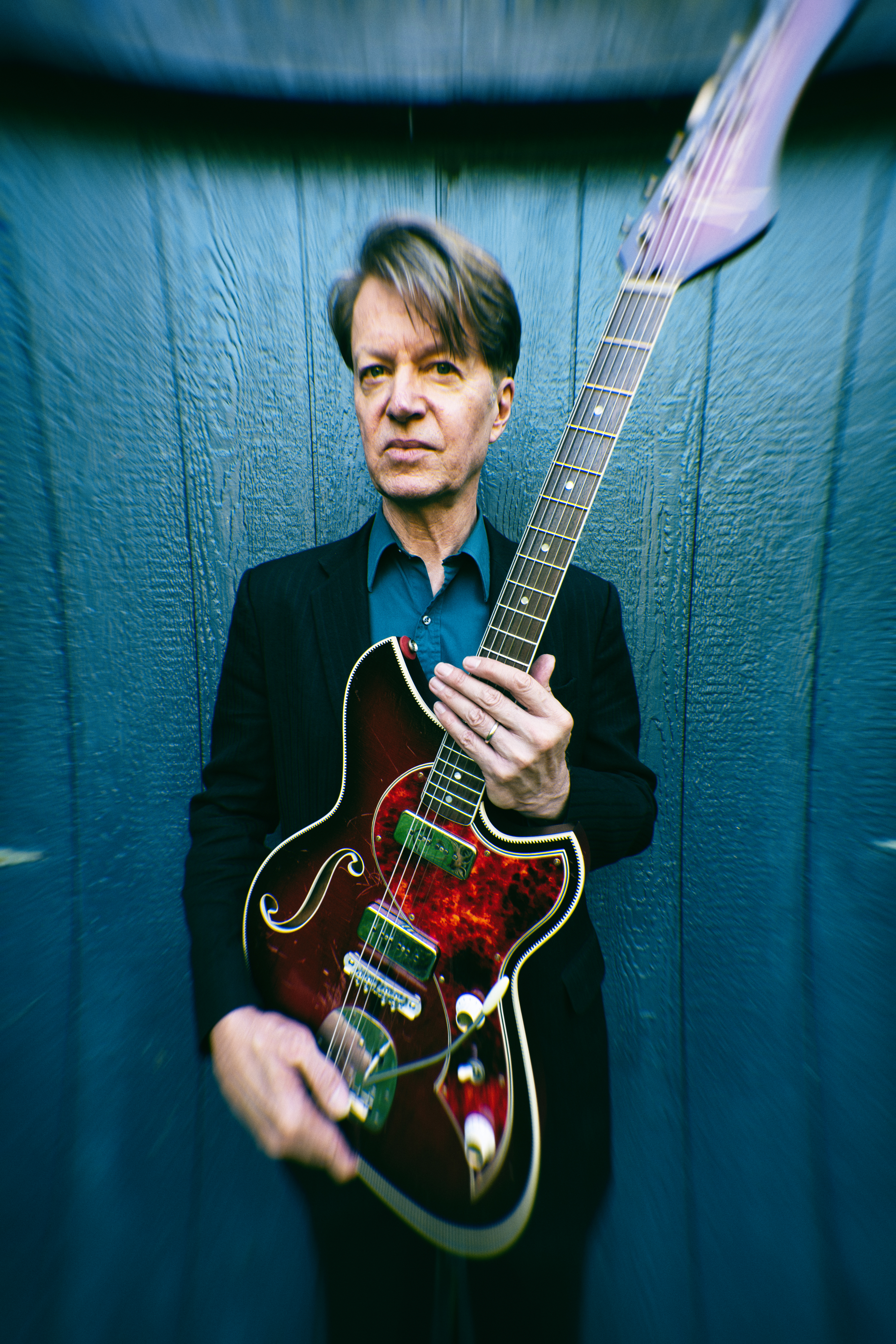 Nels Cline, holding an electric guitar upright, faces the camera, wearing a suit