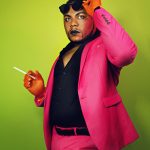 Dezi 5, against a neon green backdrop, is wearing a hot pink suit and black shirt, holding a cigarette and raising sunglasses off his eyes