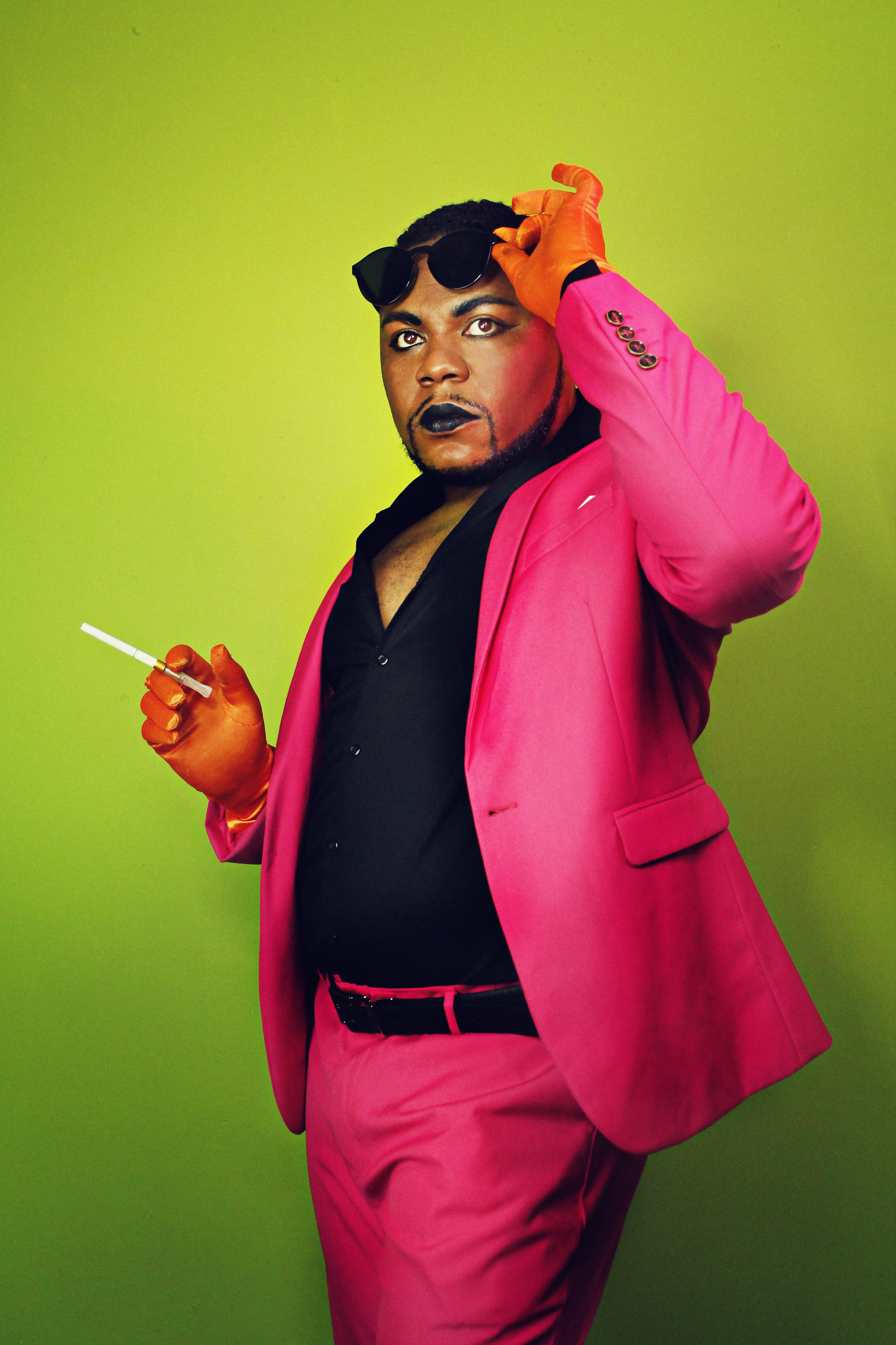 Dezi 5, against a neon green backdrop, is wearing a hot pink suit and black shirt, holding a cigarette and raising sunglasses off his eyes