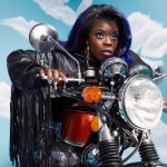 Yola, wearing a leather motorcycle jacket, is astride a motorcycle against a blue sky