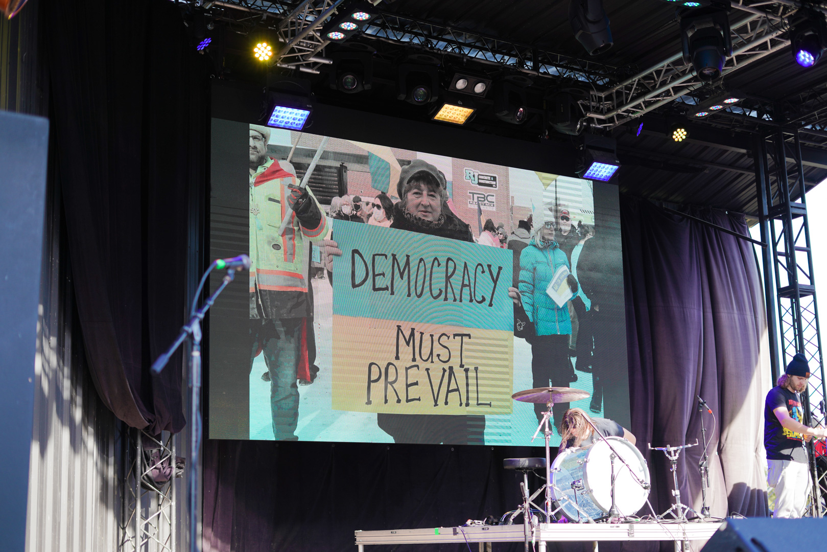 An LED screen shows a woman holding a sign that says "Democracy Must Prevail"