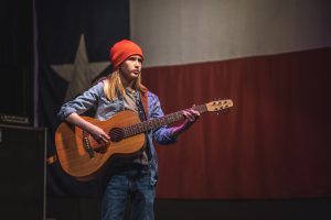 Jack Barksdale, clad in a red knit cap and blue shirt, holds an acoustic guitar in front of a large Texas state flag
