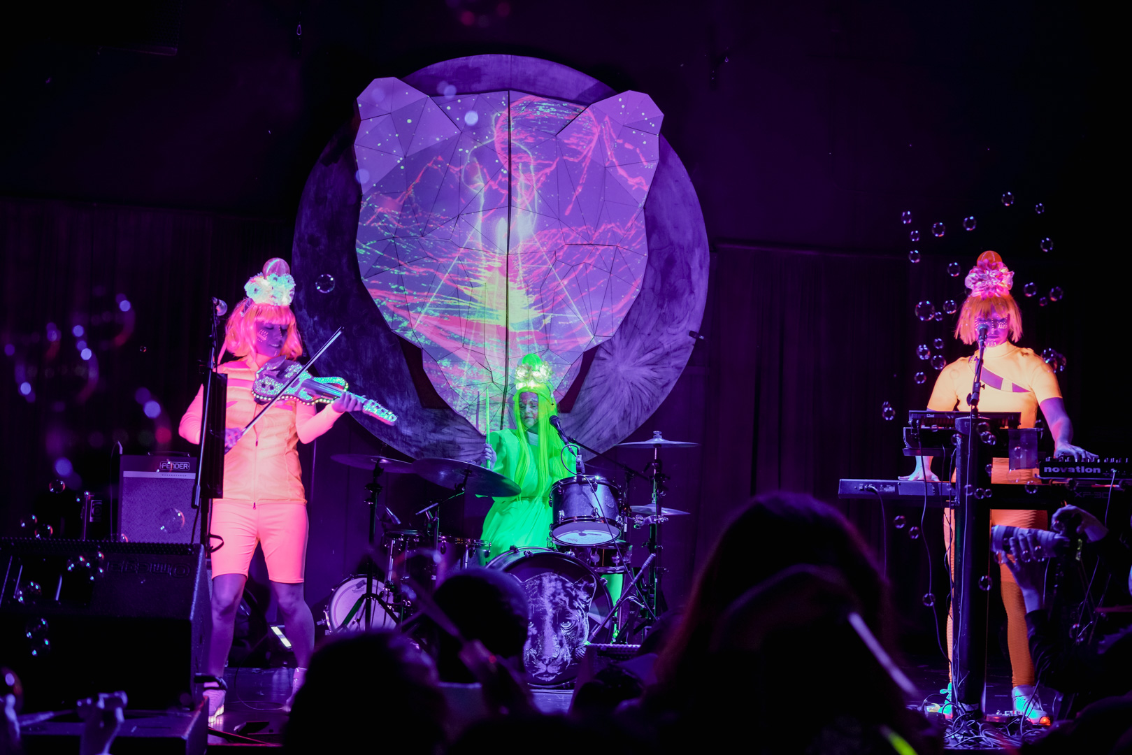 3 female musicians on stage in highlighter color outfit with an alien aesthetic play violin, keyboards and drums on stage