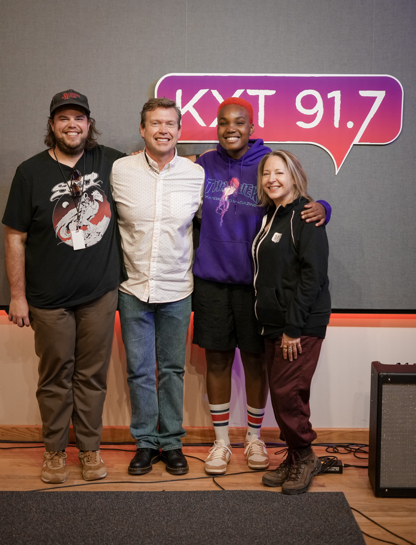 Two men and two women stand in front of "KXT91.7" sign while smiling
