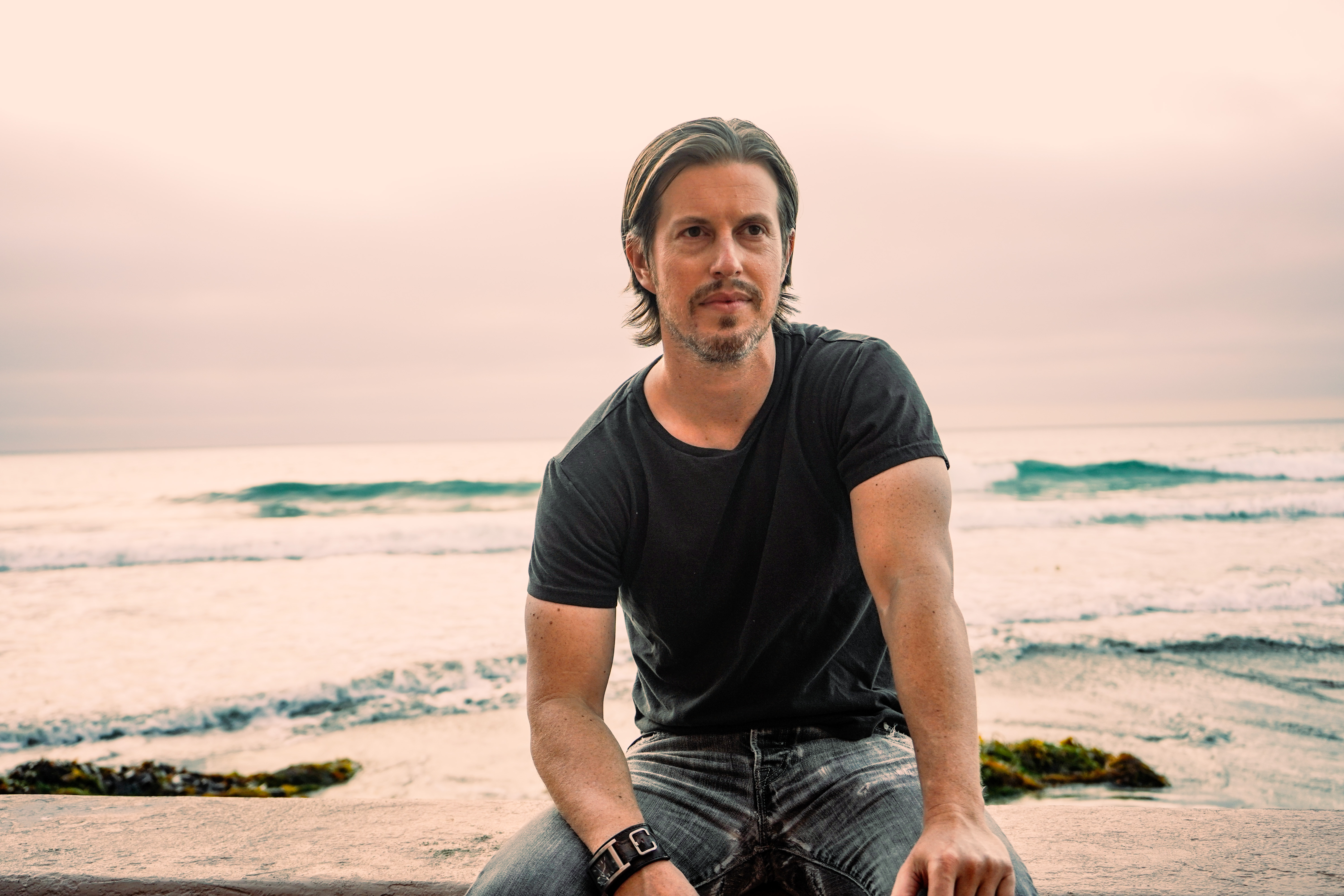 Wearing a black T-shirt and blue jeans, Barton Stanley David faces the camera, the ocean behind him