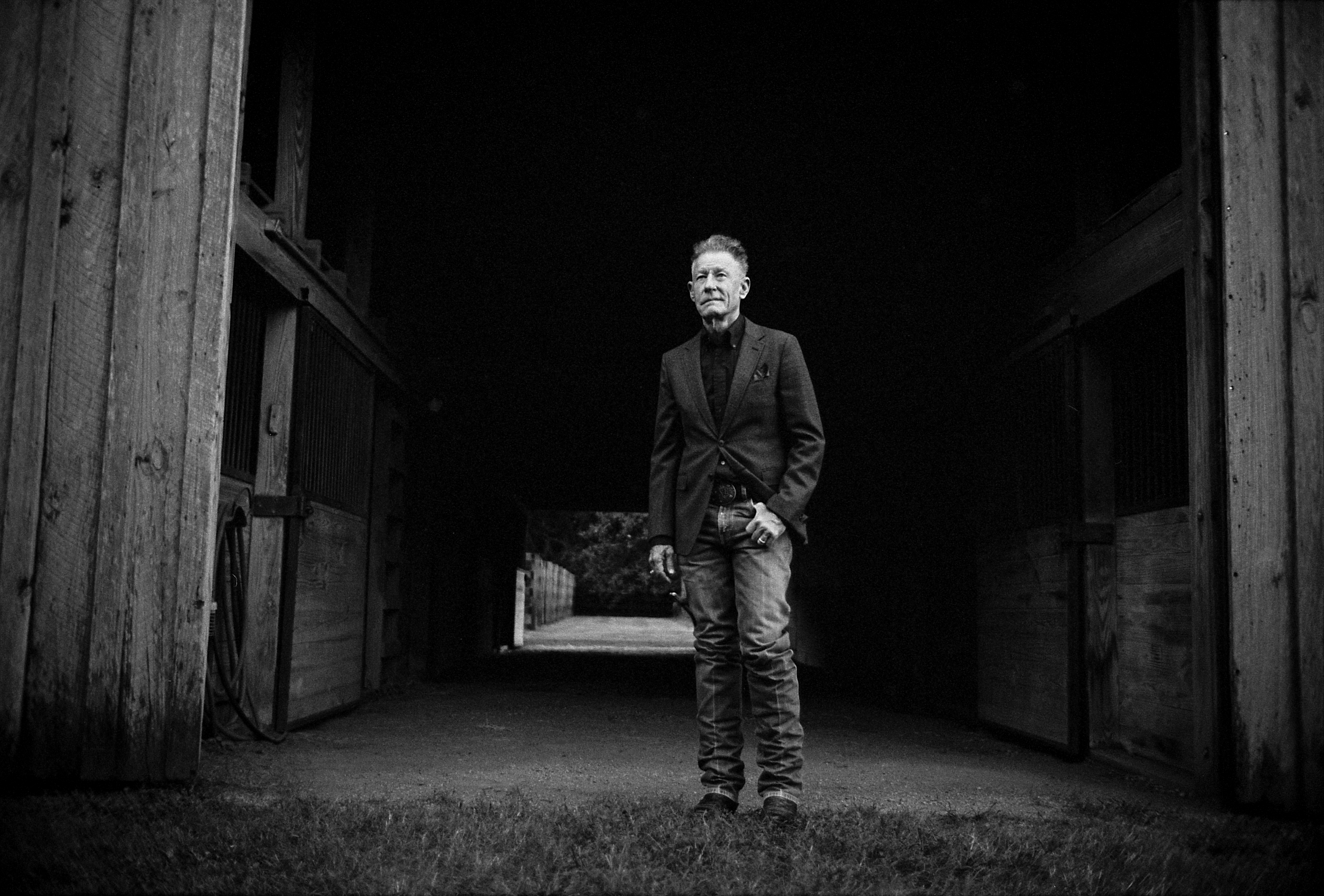Lyle Lovett, in jeans and a button-up jacket, stands inside a horse barn