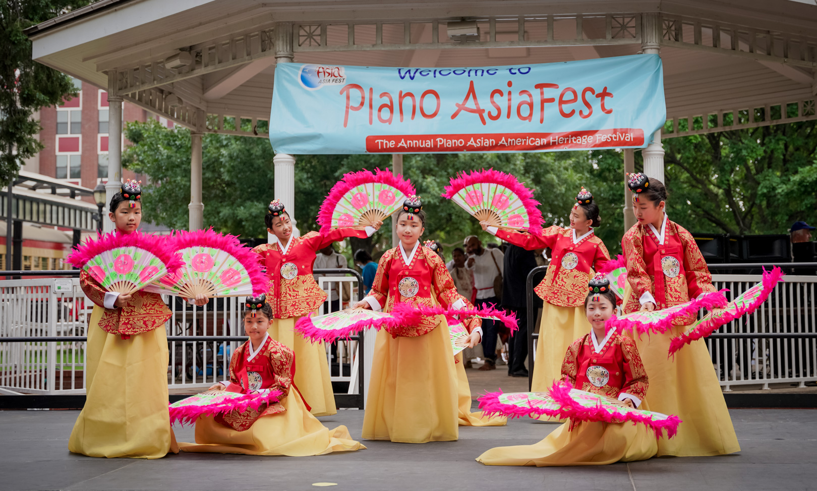 8 young girls in red and gold dresses dance with pink hand fans