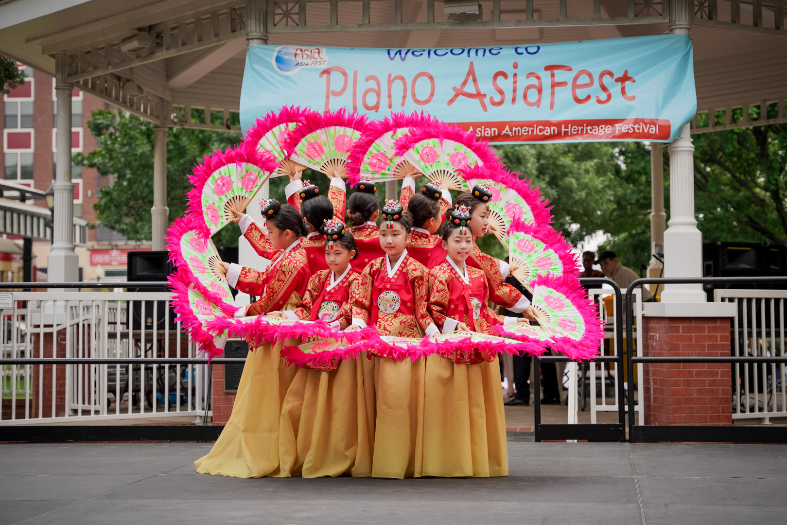 8 young girls in red and gold dresses dance with pink hand fans