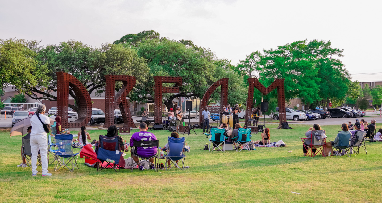 An art installation 12 ft tall with the word DREAM in a grassy park. A band plays in front of it