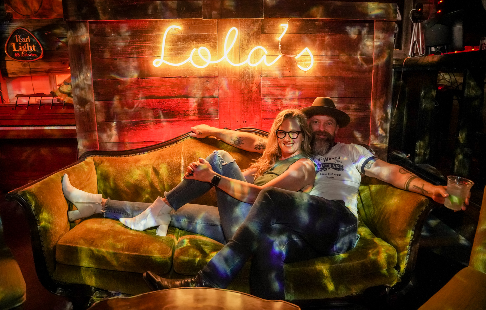 A woman and man sit on a couch with a neon sign that says "Lola's"