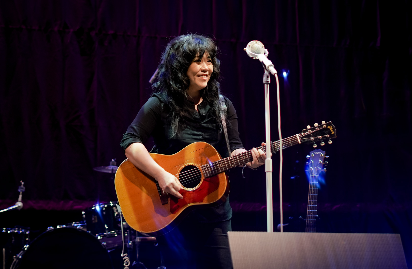 A woman smiles with a guitar on stage