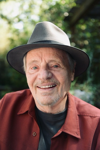 Wearing a fedora and a red shirt, Delbert McClinton faces the camera