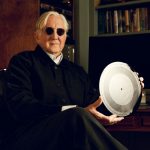 T Bone Burnett, dressed in black and wearing sunglasses, holds up a silver disc