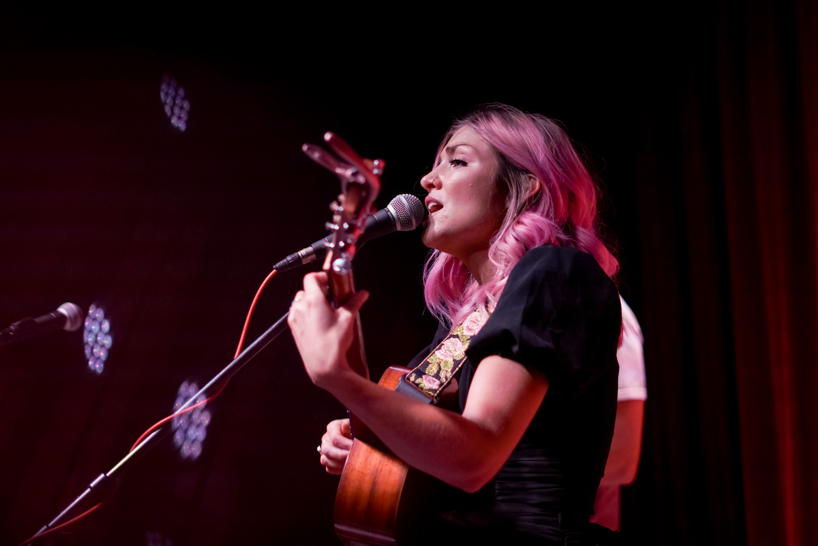 A woman with pink hair playing an acoustic guitar and sings on stage