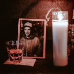 A photograph, a lit candle, and a whiskey shot sitting on a bass speaker