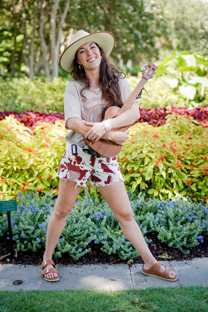 A woman smiles holding a ukulele in front of a flower garden