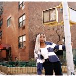 Wearing a black and white striped sweater, Hannah Jadagu stands next to a light pole on the street