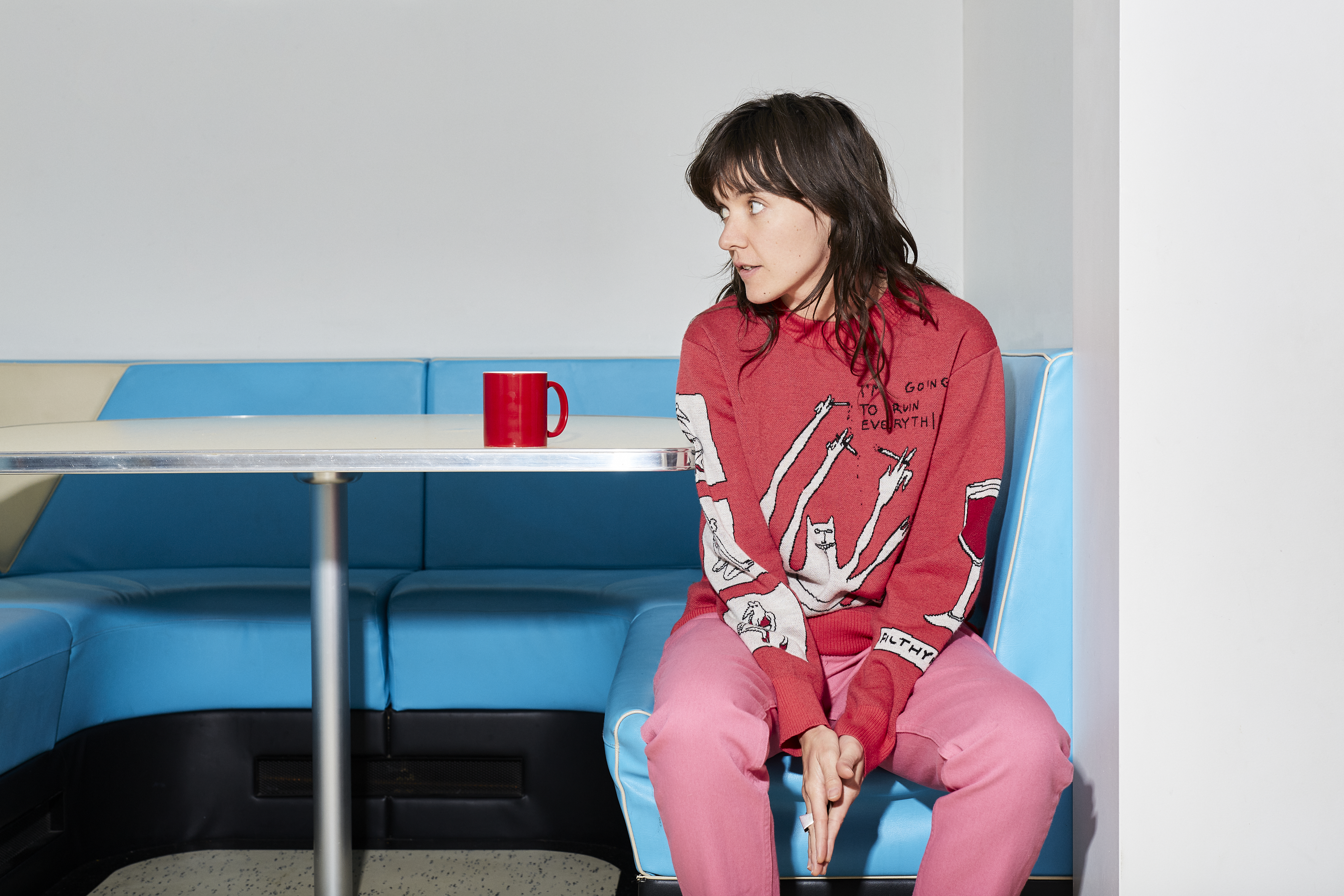 Seated in a blue booth, wearing a red shirt, Courtney Barnett looks off to the left