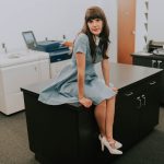 KellyMarie, wearing a dress, sits inside a office